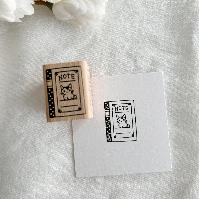 Pottering Cat rubber stamp titled 'Notebook,' featuring an adorable cat on the cover of a notebook, Japanese stationery perfect for adding a study-themed, creative touch to planners, journals, and crafts.